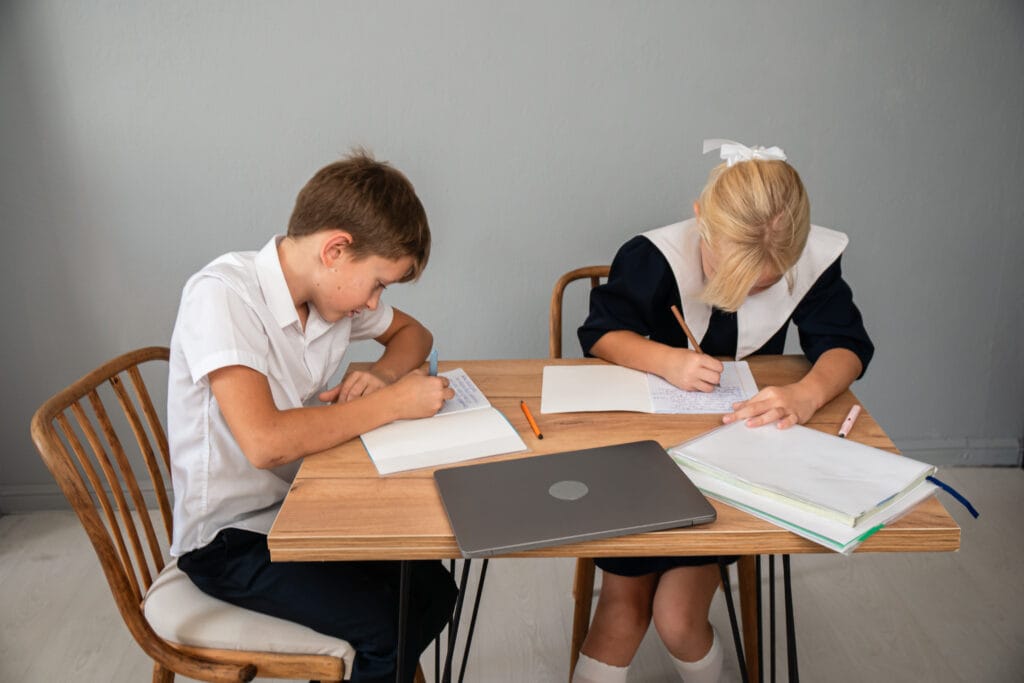 Two elementary school kids in uniforms doing homework together at a table