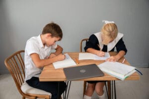Two elementary school kids in uniforms doing homework together at a table